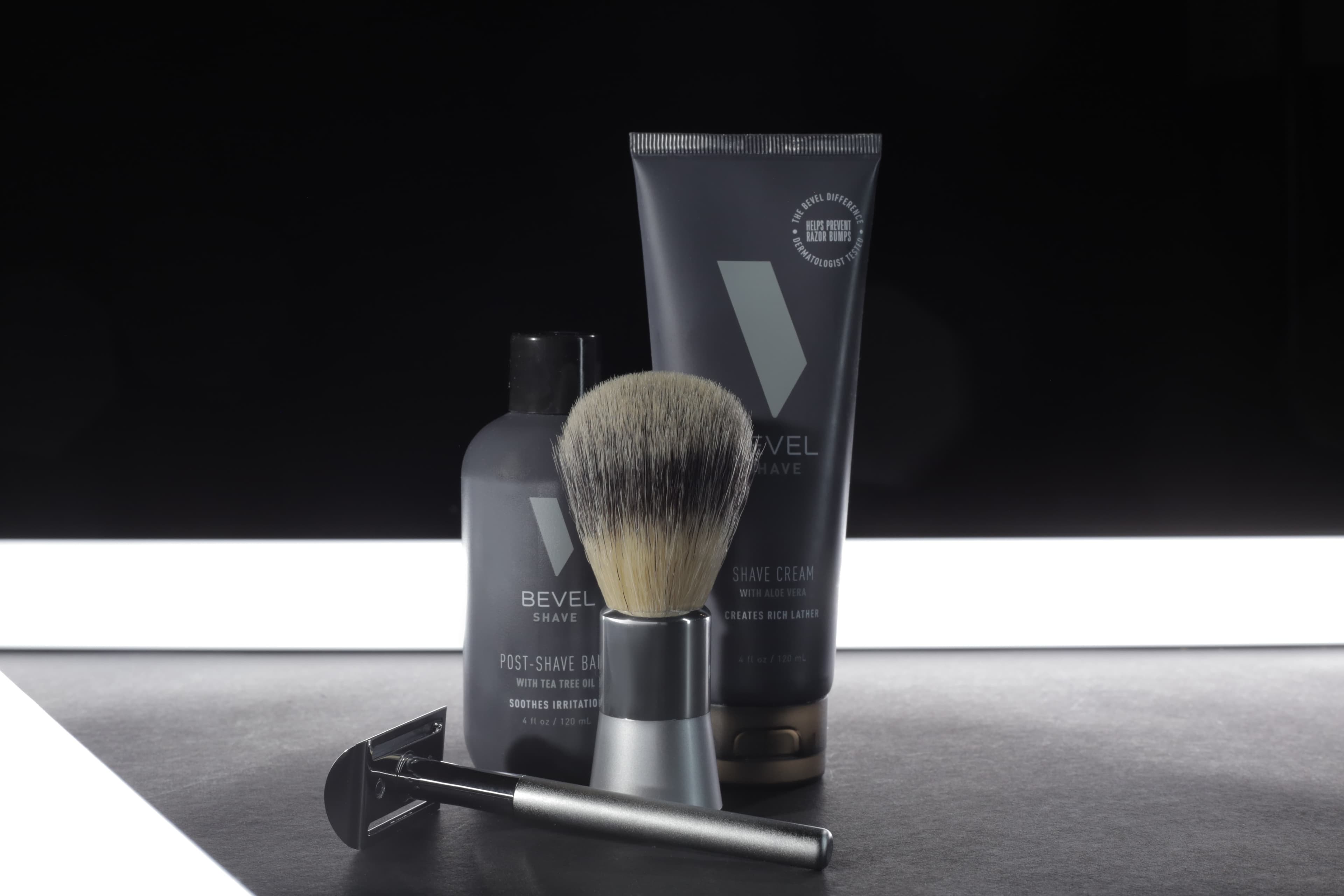 Bevel shaving products arranged on a dark surface, including a shaving brush, a post-shave balm, and a tube of shave cream, illuminated by a horizontal light strip.